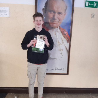 Uśmiechnięty uczeń ósmej klasy prezentuje nagrodę książkową i dyplom, w tle znajduje się obraz z Janem Pawłem II.
