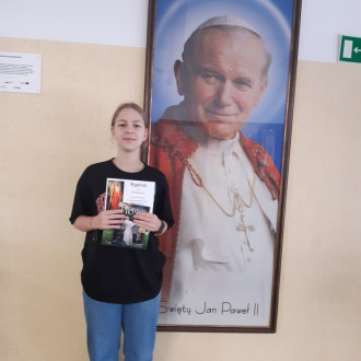 Uśmiechnięta uczennica ósmej klasy prezentuje nagrodę książkową i dyplom, w tle znajduje się obraz z Janem Pawłem II.