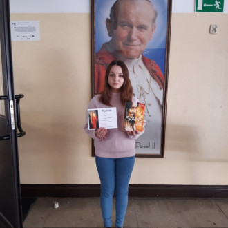 Uśmiechnięta uczennica ósmej klasy prezentuje nagrodę książkową i dyplom, w tle znajduje się obraz z Janem Pawłem II.