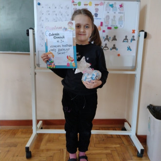 Uśmiechnięta uczennica z klasy I z dyplomem i słodkim upominkiem, w tle sala lekcyjna