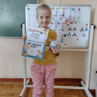 Uśmiechnięta uczennica z klasy I z dyplomem i słodkim upominkiem, w tle sala lekcyjna