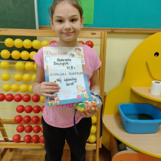 Uśmiechnięta uczennica z klasy II z dyplomem i słodkim upominkiem, w tle sala lekcyjna